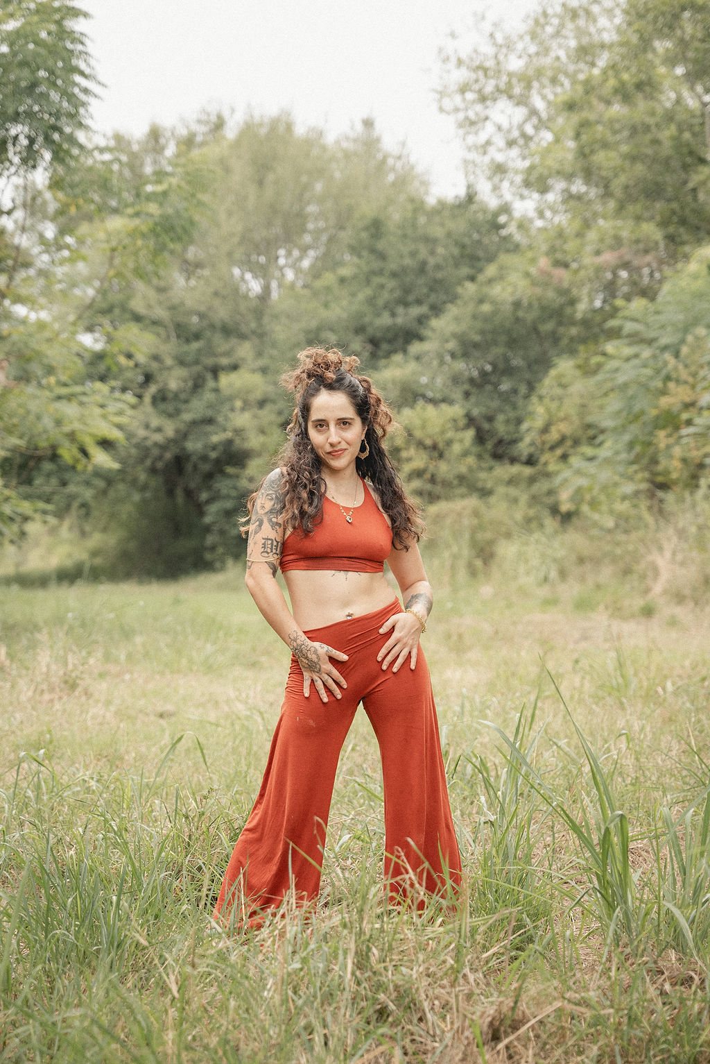 Diana L. Origel de pie en un campo, con ropa color terracota y tatuajes en brazos y manos
