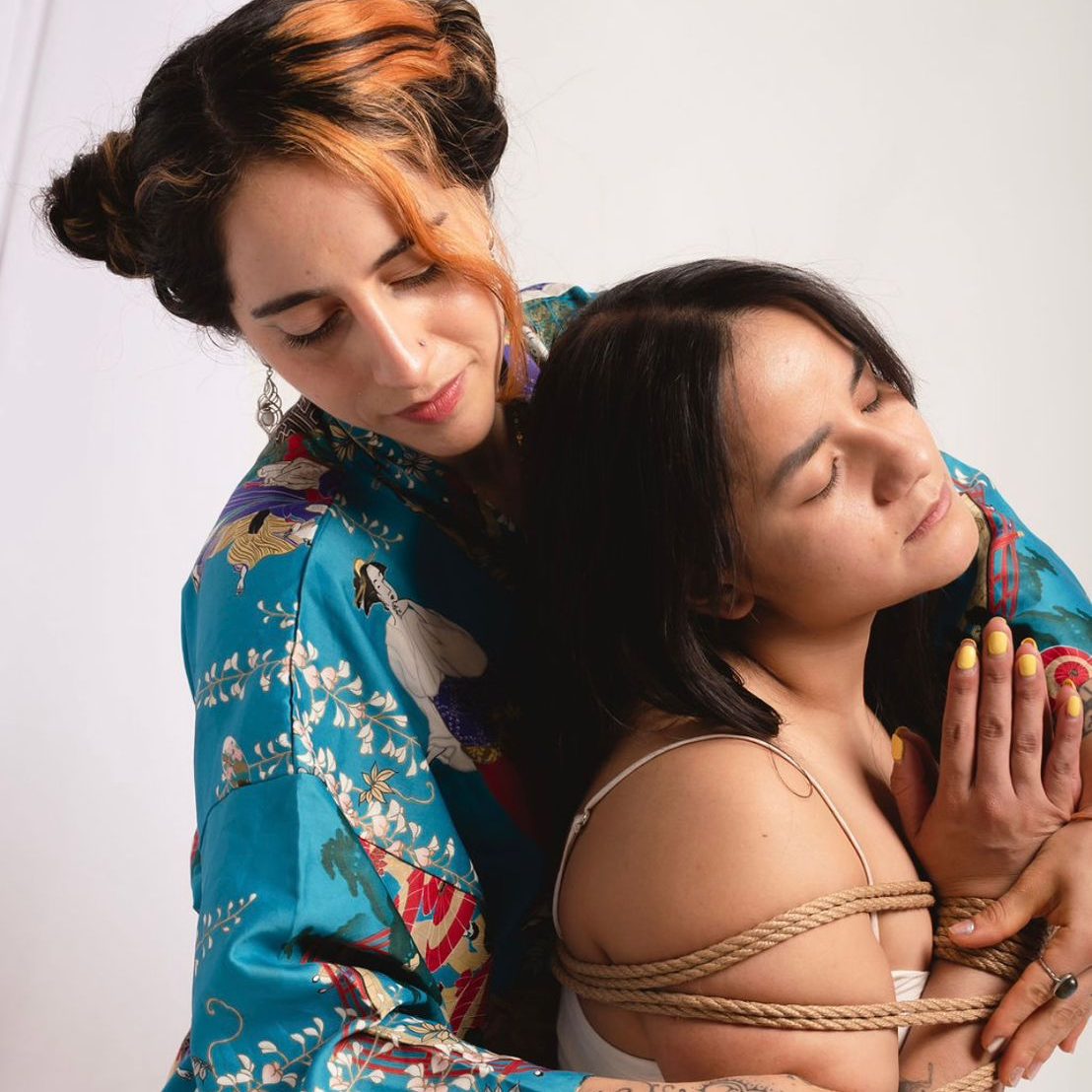 Una persona con kimono estampado abraza por la espalda a otra que tiene cuerdas de yute sobre los hombros y los ojos cerrados