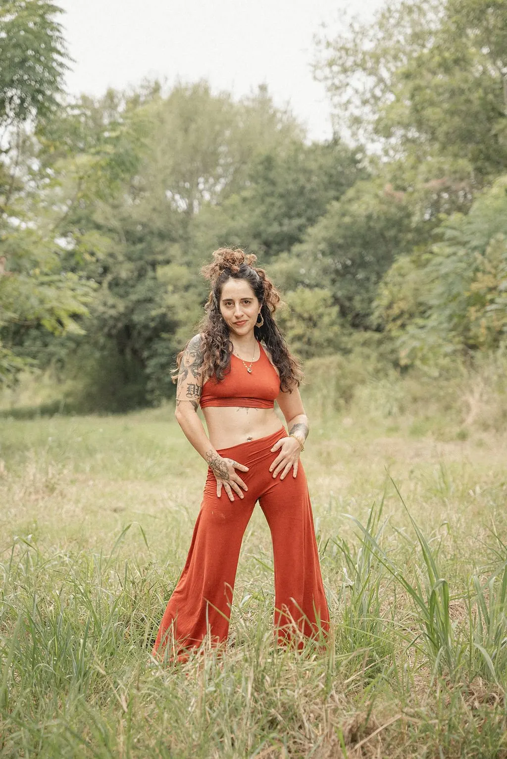Diana L. Origel de pie en un campo, con ropa color terracota y tatuajes en brazos y manos